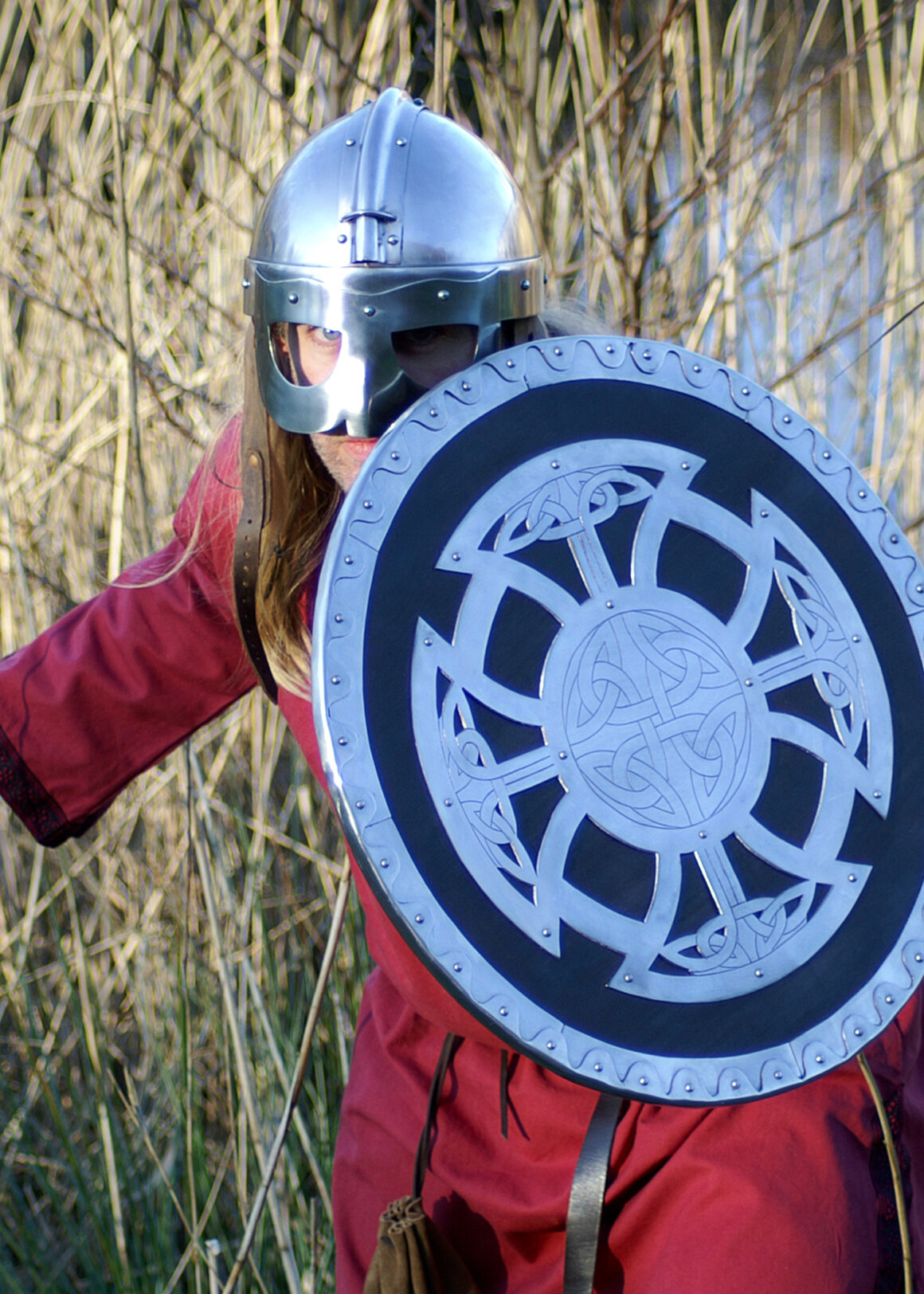ULFBERTH Wikinger Rundschild, Schaukampftauglich (Holz, Stahl, Leder) 54,5 cm Durchmesser - eine Wikingerkrieger steht mit Schild und Helm am Schilf an einem See