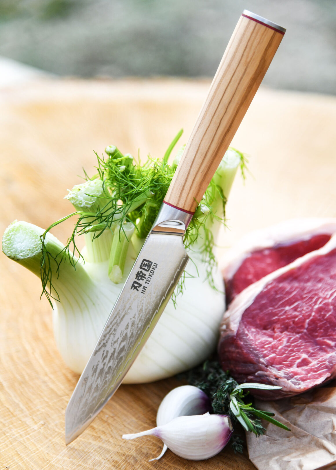 Ha Teikoku Allzweckmesser Damaststahl Olivenholz Griff - Messer lehnt an Fenchel, auf einem Holzbrett, mit frischen Kräutern, Knoblauch und einem Rindersteak