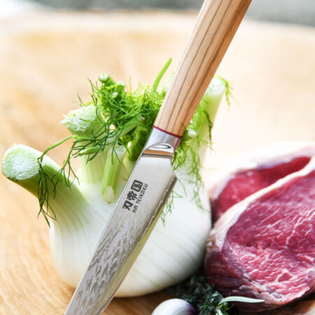 Battle Merchant Allzweckmesser Damaststahl Olivenholz Griff - Messer lehnt an Fenchel, auf einem Holzbrett, mit frischen Kräutern, Knoblauch und einem Rindersteak