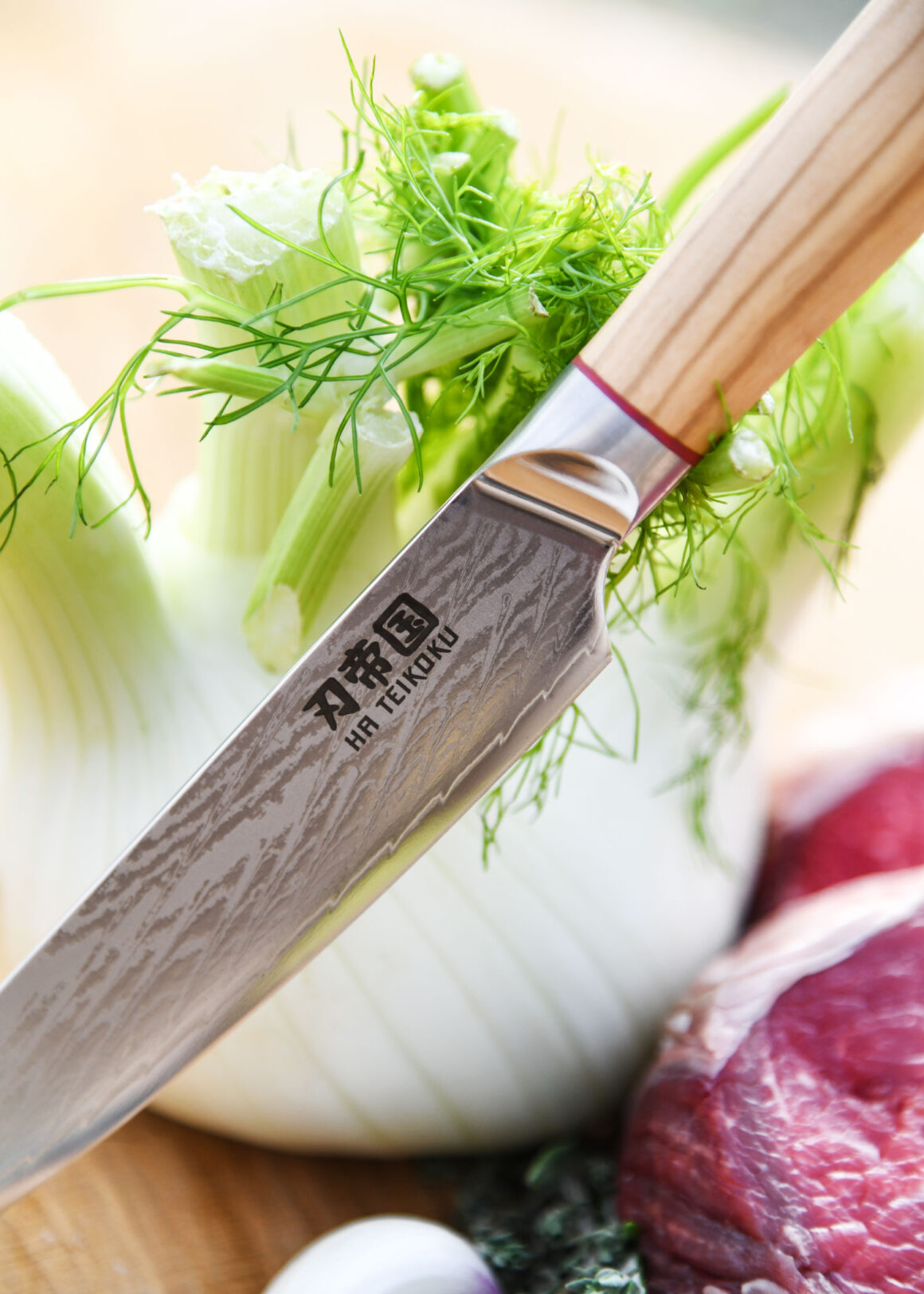 Ha Teikoku Allzweckmesser Damaststahl Olivenholz Griff - Nahaufnahme Messer lehnt an Fenchel, auf einem Holzbrett, mit frischen Kräutern, Knoblauch und einem Rindersteak