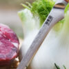 Ha Teikoku Ausbeinmesser, Damaststahl, Olivenholz - Nahaufnahme Klinge lehnt an ein Fenchel an, auf einer Holzscheibe mit einem Rindersteak und Kräutern