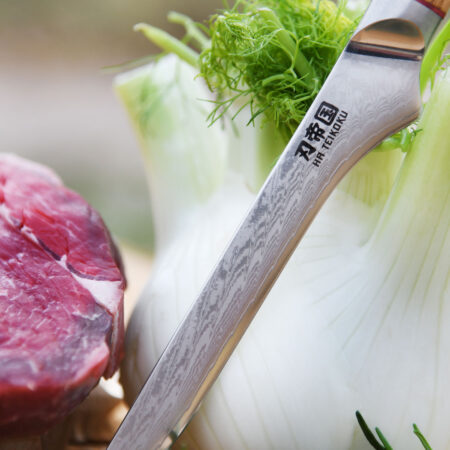 Ha Teikoku Ausbeinmesser, Damaststahl, Olivenholz - Nahaufnahme Klinge lehnt an ein Fenchel an, auf einer Holzscheibe mit einem Rindersteak und Kräutern
