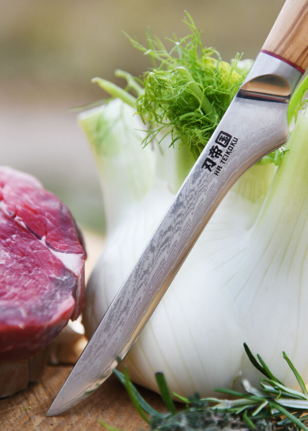 Ha Teikoku Ausbeinmesser, Damaststahl, Olivenholz - Nahaufnahme Klinge lehnt an ein Fenchel an, auf einer Holzscheibe mit einem Rindersteak und Kräutern