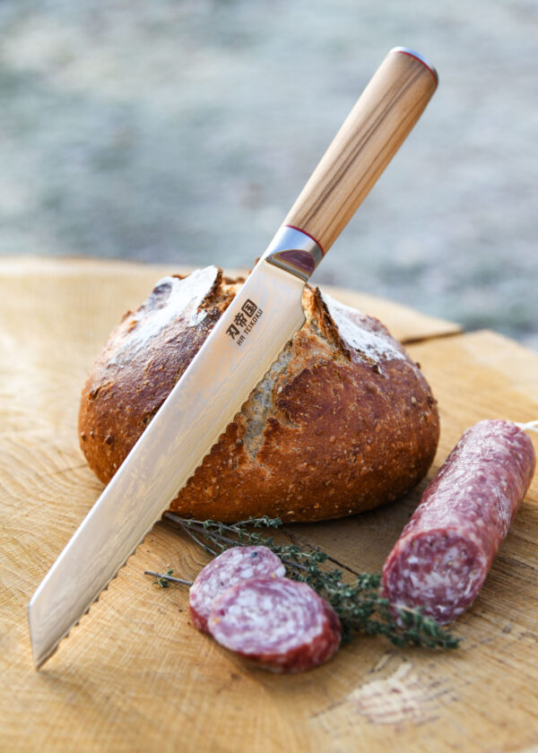 Ha Teikoku Brotmesser Damaststahl Olivenholz - Klinge lehnt an ein Brotlaib und Holzscheibe an, mit einer aufgeschinittenen Salamistange und Thymian daneben