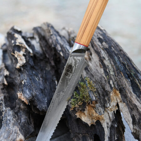 Ha Teikoku Brotmesser Damaststahl Olivenholz - Klinge lehnt an ein altes graues Stück Holz und einem Holztisch