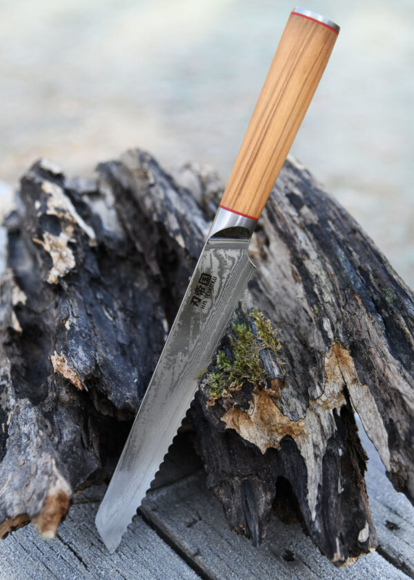 Ha Teikoku Brotmesser Damaststahl Olivenholz - Klinge lehnt an ein altes graues Stück Holz und einem Holztisch