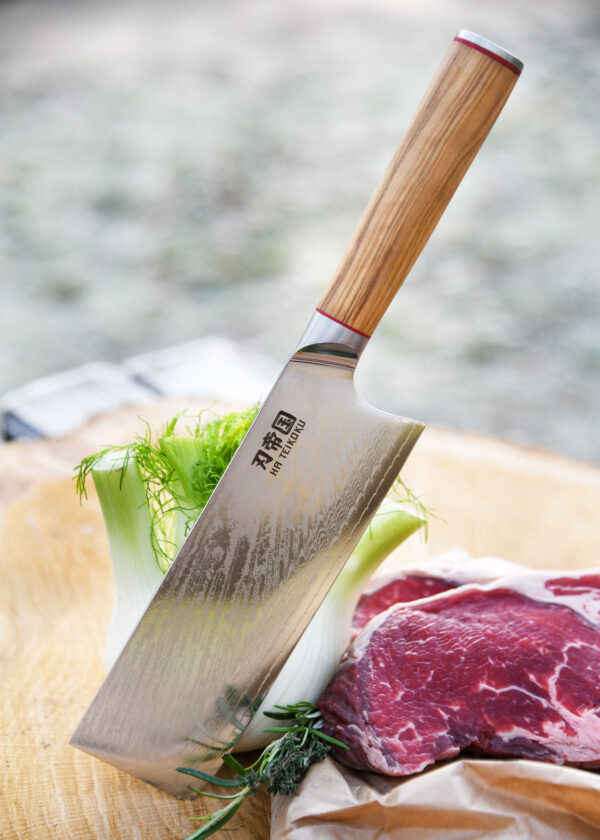 Ha Teikoku Hackmesser, Damaststahl Olivenholzgriff - Seitenansicht, Beil steckt in einem Holzbrett an ein Fenchel gelehnt, mit 2 Stück frisch geschnittenen Rindfleischsteaks