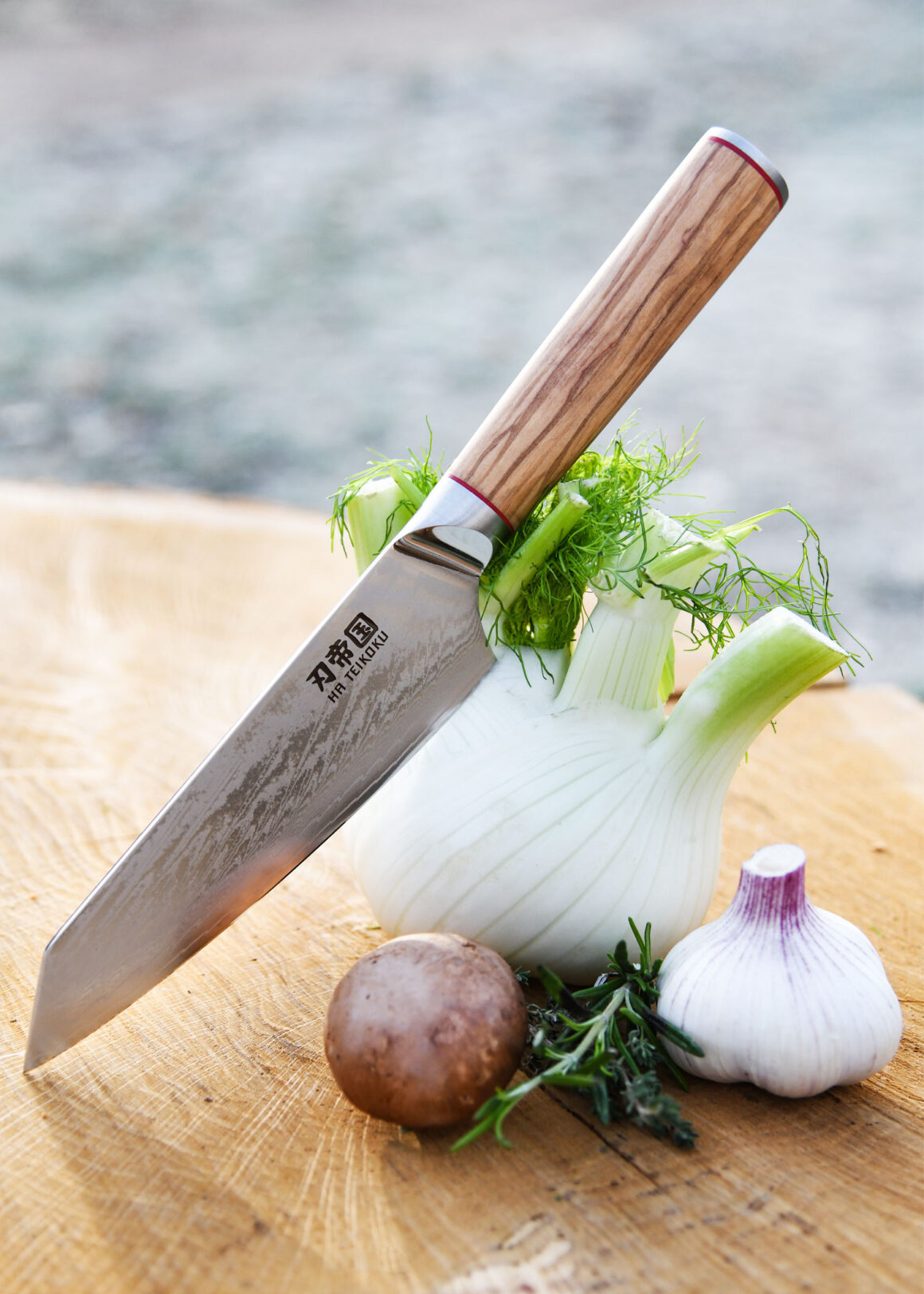 Ha Teikoku Kochmesser, 20 cm Klingenlänge, Damaststahl Olivenholz - Messer lehnt an einem Fenchel, auf einem Holzbrett mit frischem Knoblauch, Kräutern und einer Kartoffel