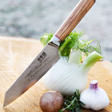 Ha Teikoku Kochmesser, 20 cm Klingenlänge, Damaststahl Olivenholz - Messer lehnt an einem Fenchel, auf einem Holzbrett mit frischem Knoblauch, Kräutern und einer Kartoffel