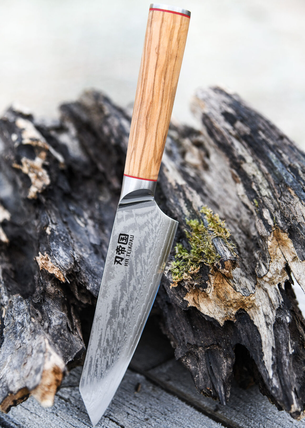 Ha Teikoku Kochmesser, 20 cm Klingenlänge, Damaststahl Olivenholz - Messer lehnt an ein altes graues Holzstück, auf einem Holztisch