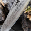 Ha Teikoku Kochmesser, 20 cm Klingenlänge, Damaststahl Olivenholz - Nahaufnahme Klinge lehnt an ein altes graues Holzstück, auf einem Holztisch
