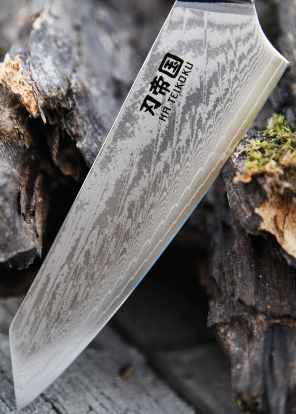 Ha Teikoku Kochmesser, 20 cm Klingenlänge, Damaststahl Olivenholz - Nahaufnahme Klinge lehnt an ein altes graues Holzstück, auf einem Holztisch