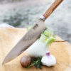 Ha Teikoku Kochmesser 24 cm Klingenlänge Damaststahl - Messer liegt an einem Fenchel mit Knoblauch, Kartoffel und Kräutern an und zeigt mit der Messerspitze auf eine Baumscheibe, steiniger Hintergrund verschwommen