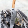 Ha Teikoku Kochmesser 24 cm Klingenlänge Damaststahl - Messer lehnt an einen grauen holzstamm mit der Klinge auf einem holztisch, der hintergrund verschwommen
