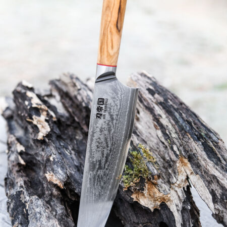 Ha Teikoku Kochmesser 24 cm Klingenlänge Damaststahl - Messer lehnt an einen grauen holzstamm mit der Klinge auf einem holztisch, der hintergrund verschwommen