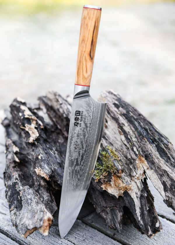 Ha Teikoku Kochmesser 24 cm Klingenlänge Damaststahl - Messer lehnt an einen grauen holzstamm mit der Klinge auf einem holztisch, der hintergrund verschwommen