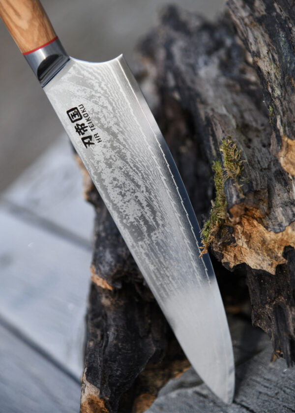 Ha Teikoku Kochmesser 24 cm Klingenlänge Damaststahl - Klinge lehnt an einen grauen holzstamm, der hintergrund verschwommen
