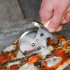 Battle Merchant Pizzaschneider Pizzaroller Wikinger Schwert Edelstahl, Eschenholz, Stahl - Hand hält den Pizzaroller und schneidet eine Pizza auf einem Marmortisch, Ansicht von leicht oben
