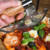 Battle Merchant Pizzaschneider Wiegemesser Wikinger Bartaxt für Pizza und Kräuter Edelstahl, Eschenholz - Hand hält Messer am Griff und schneidet in eine Pizza