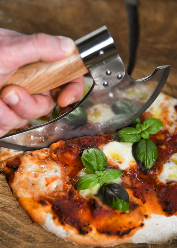 Battle Merchant Pizzaschneider Wiegemesser Wikinger Bartaxt für Pizza und Kräuter Edelstahl, Eschenholz - Hand hält Messer am Griff und schneidet in eine Pizza