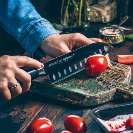 Samura MO-V Santoku Messer mit Kullenschliff 7.0 Zoll, 180 mm schwarzer Griff - Person schneidet gerade Tomaten mit dem Messer auf einem alten Holzbrett