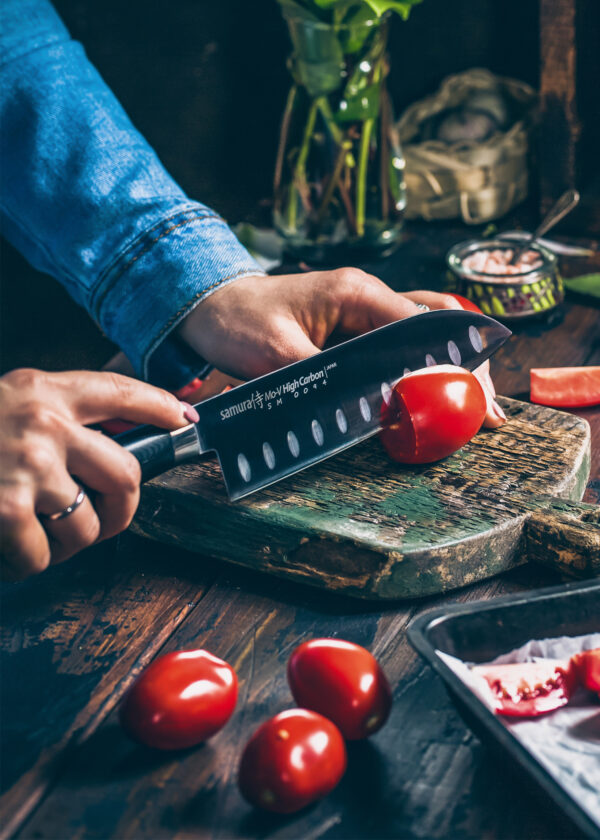 Samura MO-V Santoku Messer mit Kullenschliff 7.0 Zoll, 180 mm schwarzer Griff - Person schneidet gerade Tomaten mit dem Messer auf einem alten Holzbrett
