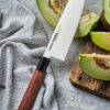 Samura Okinawa Santoku Küchenmesser 175 mm braun-schwarzer Griff - Klinge liegt auf einer aufgeschnittenen Melone und der Griff auf einem Geschirrtuch auf einem Steinboden