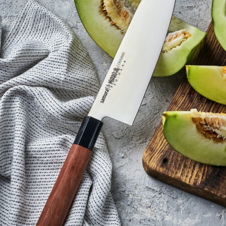 Samura Okinawa Santoku Küchenmesser 175 mm braun-schwarzer Griff - Klinge liegt auf einer aufgeschnittenen Melone und der Griff auf einem Geschirrtuch auf einem Steinboden