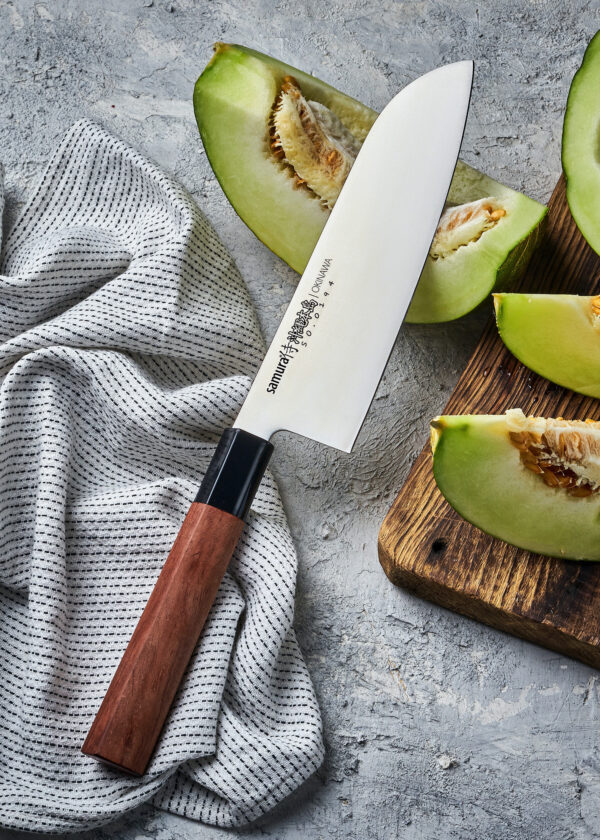 Samura Okinawa Santoku Küchenmesser 175 mm braun-schwarzer Griff - Klinge liegt auf einer aufgeschnittenen Melone und der Griff auf einem Geschirrtuch auf einem Steinboden