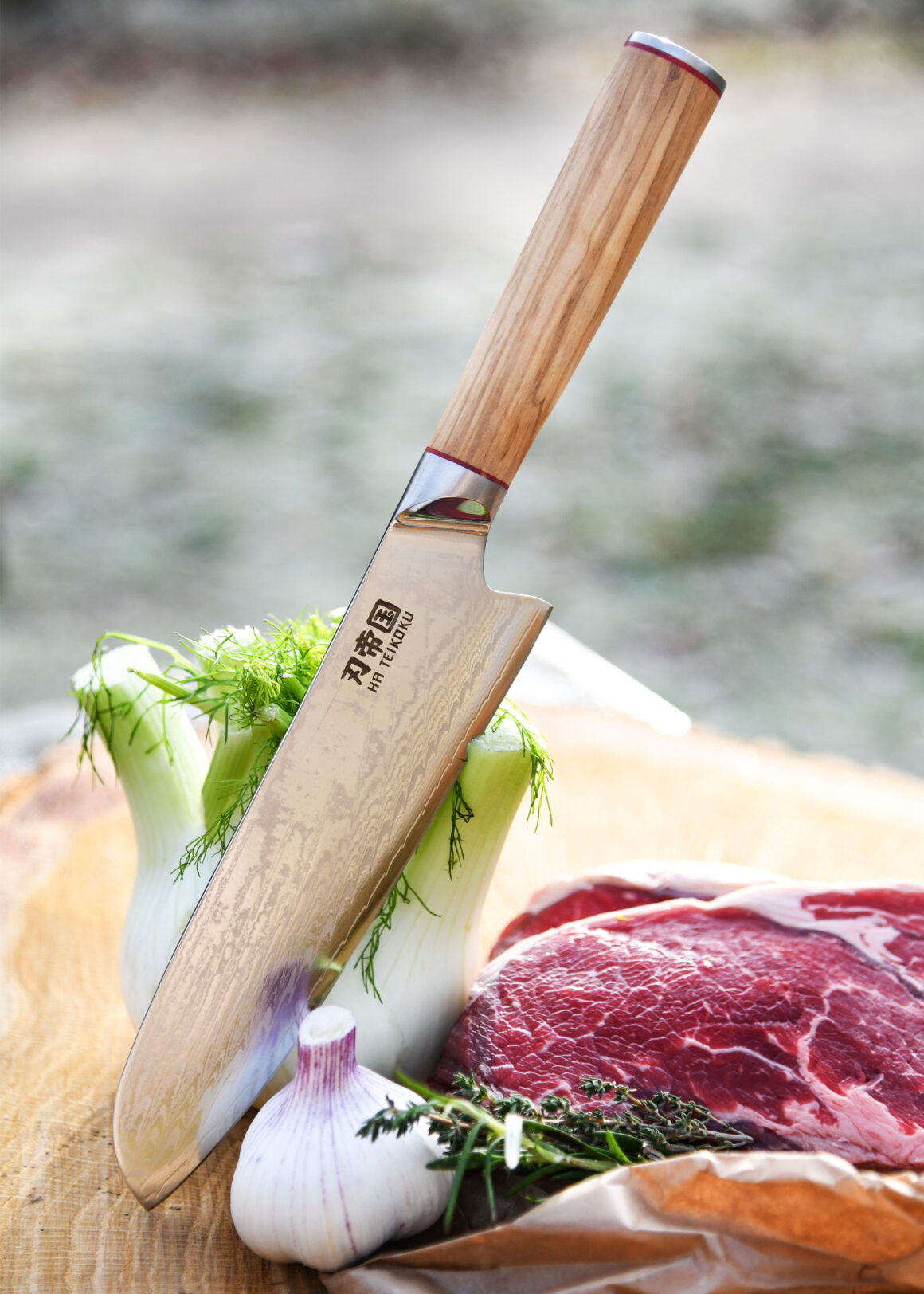 Ha Teikoku Santoku Damaststahl Olivenholz Griff - Klinge lehnt an einen Fenchel, auf einer Holzscheibe, mit frischem Knoblauch, einem Steak und ein paar Kräutern