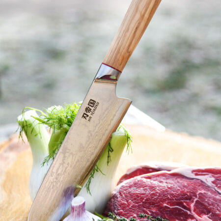Ha Teikoku Santoku Damaststahl Olivenholz Griff - Klinge lehnt an einen Fenchel, auf einer Holzscheibe, mit frischem Knoblauch, einem Steak und ein paar Kräutern