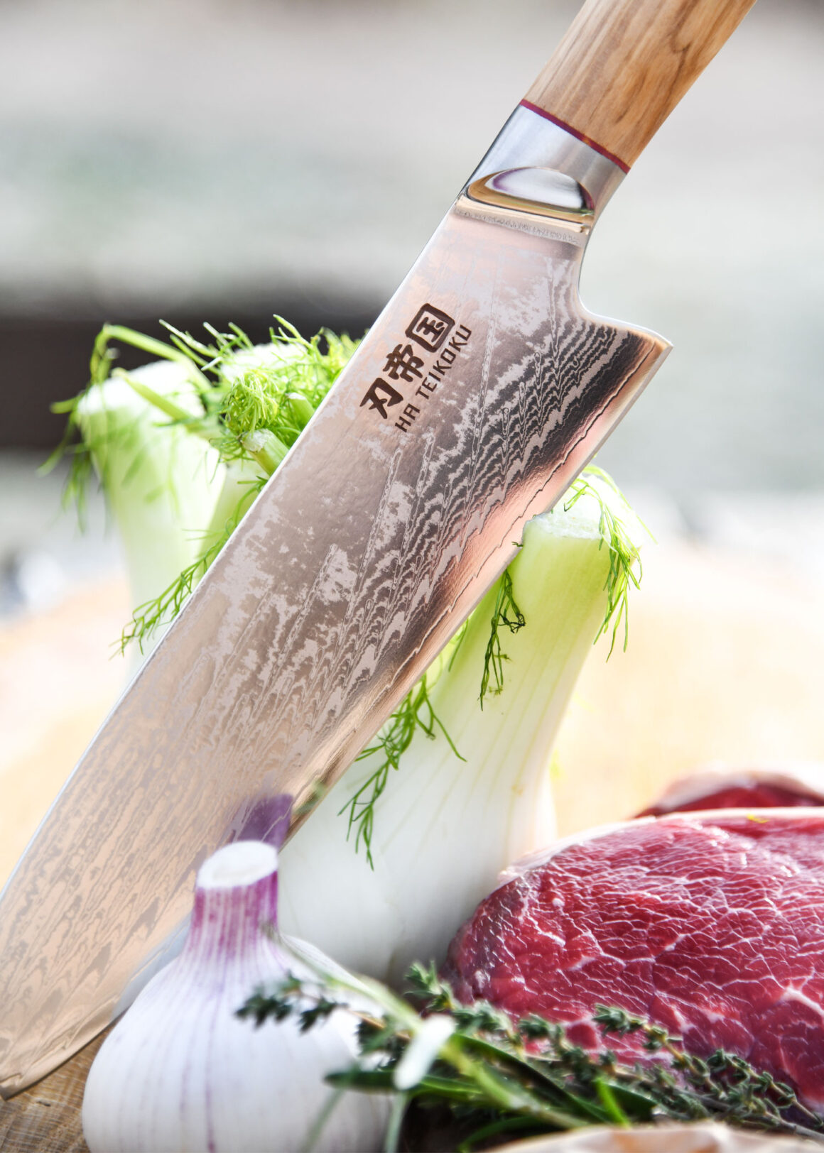 Ha Teikoku Santoku Damaststahl Olivenholz Griff - Nahaufnahme Klinge lehnt an einen Fenchel, mit frischem Knoblauch, einem Steak und ein paar Kräutern