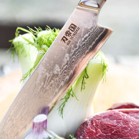 Ha Teikoku Santoku Damaststahl Olivenholz Griff - Nahaufnahme Klinge lehnt an einen Fenchel, mit frischem Knoblauch, einem Steak und ein paar Kräutern